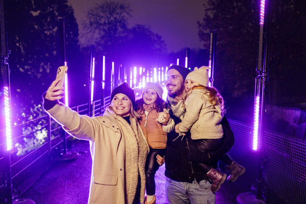 Eine vierköpfige Familie macht nachts ein Selfie auf einem Gehweg. Um sie herum leuchten senkrechte, lilafarbene Lichter.