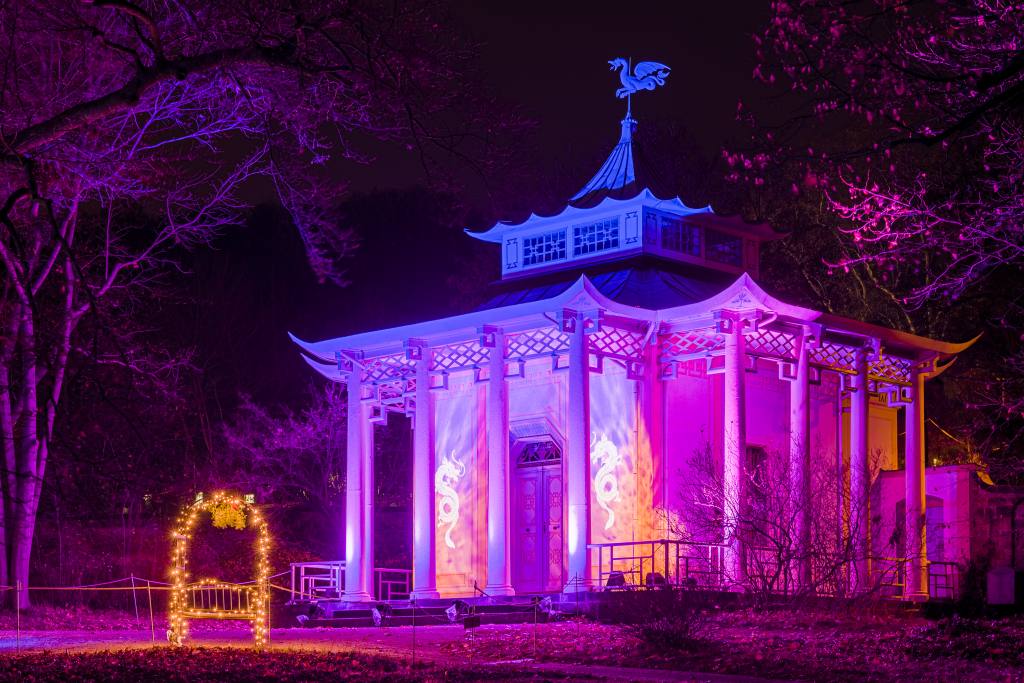 Ein kleiner Pavillon ist nachts in pinkem und lilafarbenem Licht beleuchtet. Davor steht ein beleuchteter Torbogen. Im Hintergrund befinden sich Bäume.