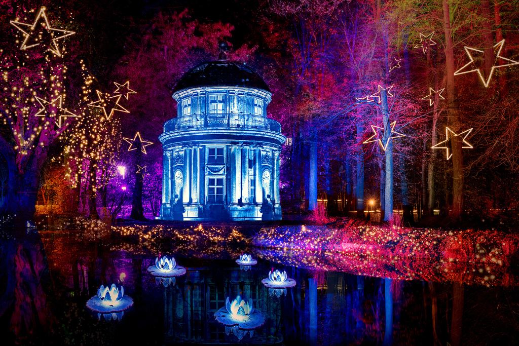 Ein rundes Gebäude ist nachts blau beleuchtet. Es steht an einem Teich. Bäume umgeben das Gebäude. Sternförmige Leuchten sind in der Umgebung verteilt. A round building is illuminated in blue at night. It stands by a pond. Trees surround the building. Star-shaped lights are distributed in the surroundings.