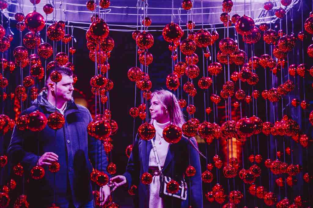 Ein Mann und eine Frau stehen unter hängenden roten Kugeln. Sie schauen sich an und lächeln. Lila Licht leuchtet auf die Szene. A man and a woman stand under hanging red balls. They look at each other and smile. Purple light shines on the scene.
