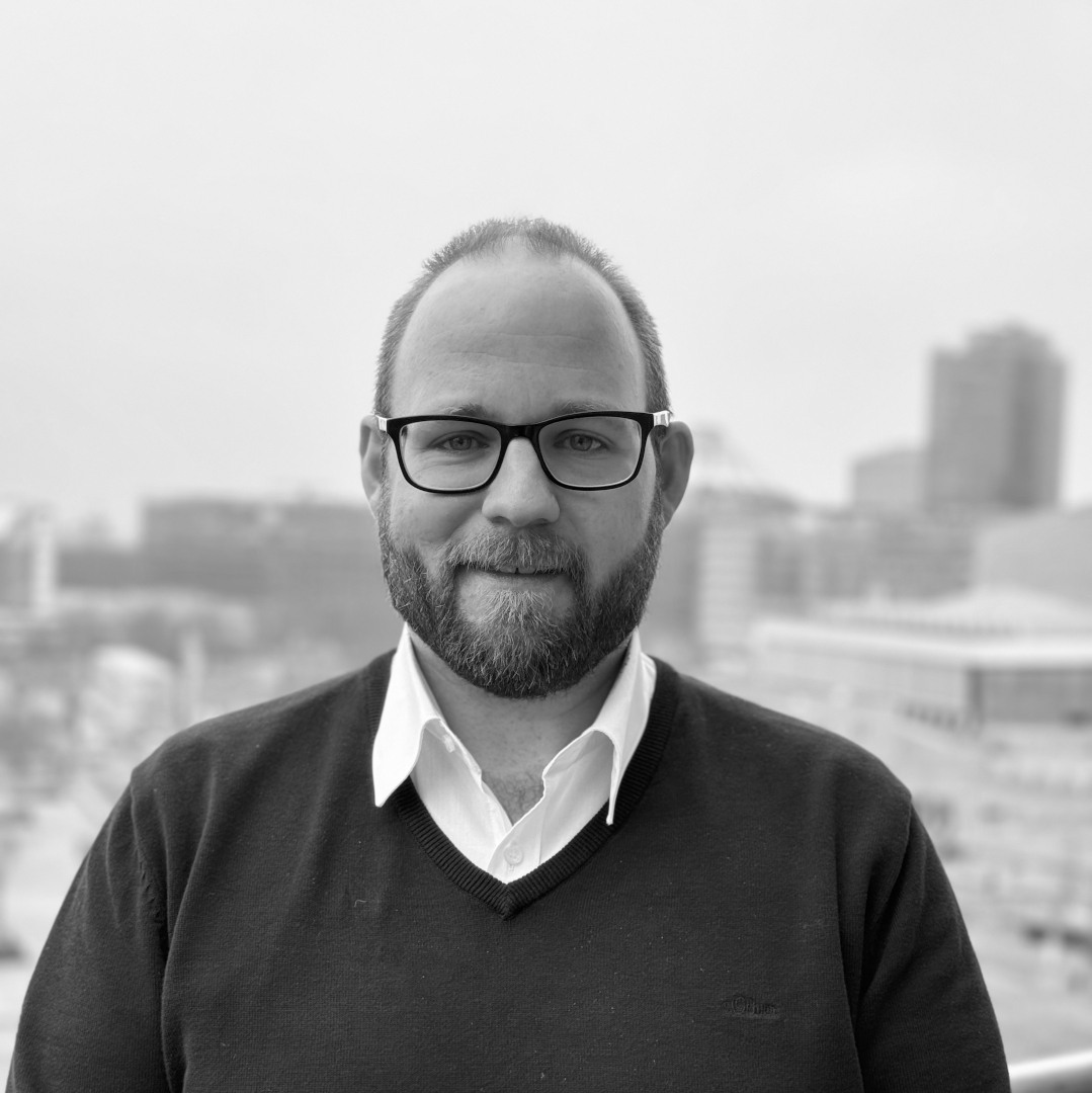 Ein Mann mit Brille und Bart steht im Freien. Er trägt ein weißes Hemd und einen dunklen Pullover. Im Hintergrund ist eine Stadtlandschaft zu sehen.