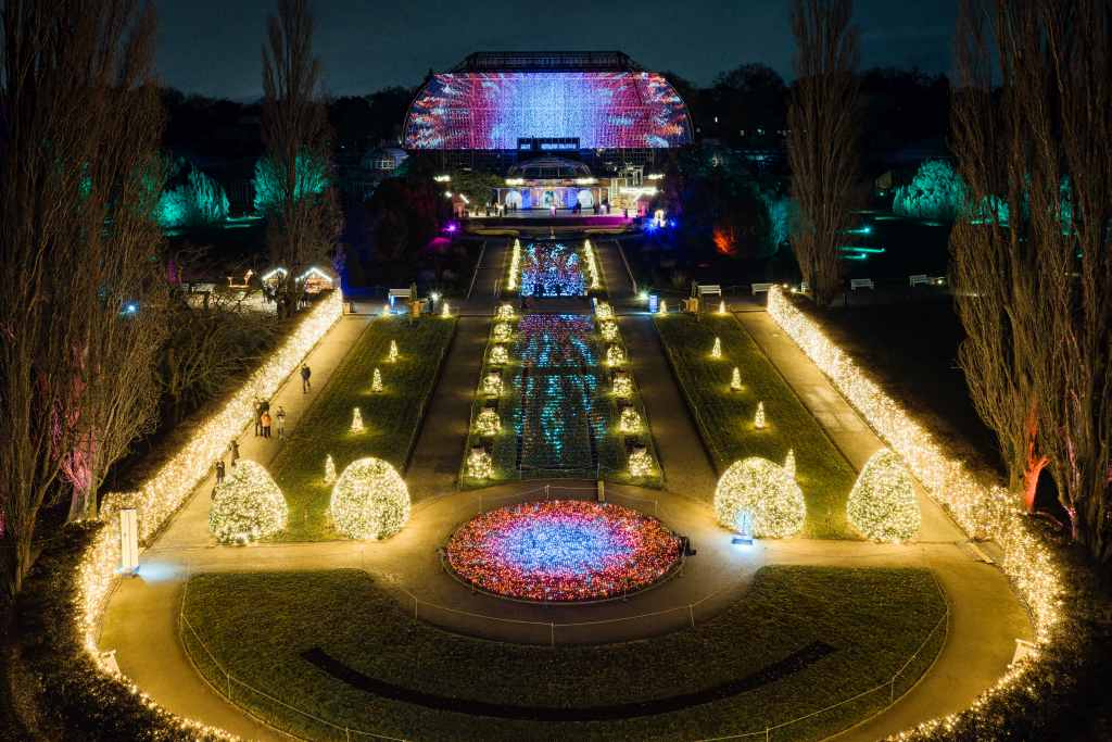 Ein großer Garten ist mit Lichtern geschmückt. Im Hintergrund steht ein gläsernes Gebäude, das bunt beleuchtet ist.