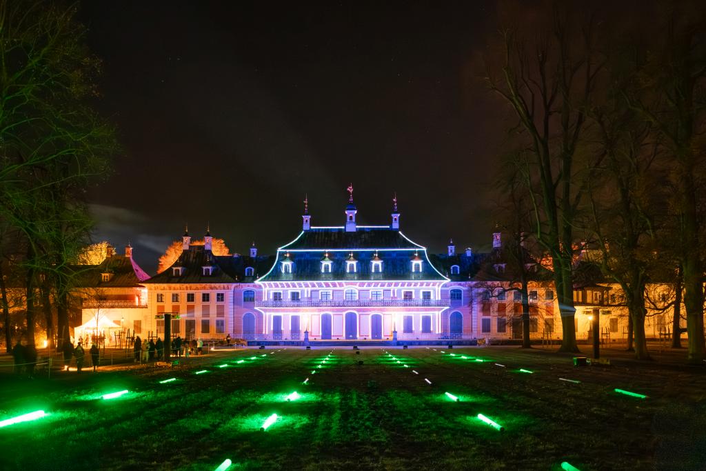 Ein großes, historisches Gebäude ist nachts bunt beleuchtet. Auf dem Rasen davor sind grüne Lichter angeordnet.