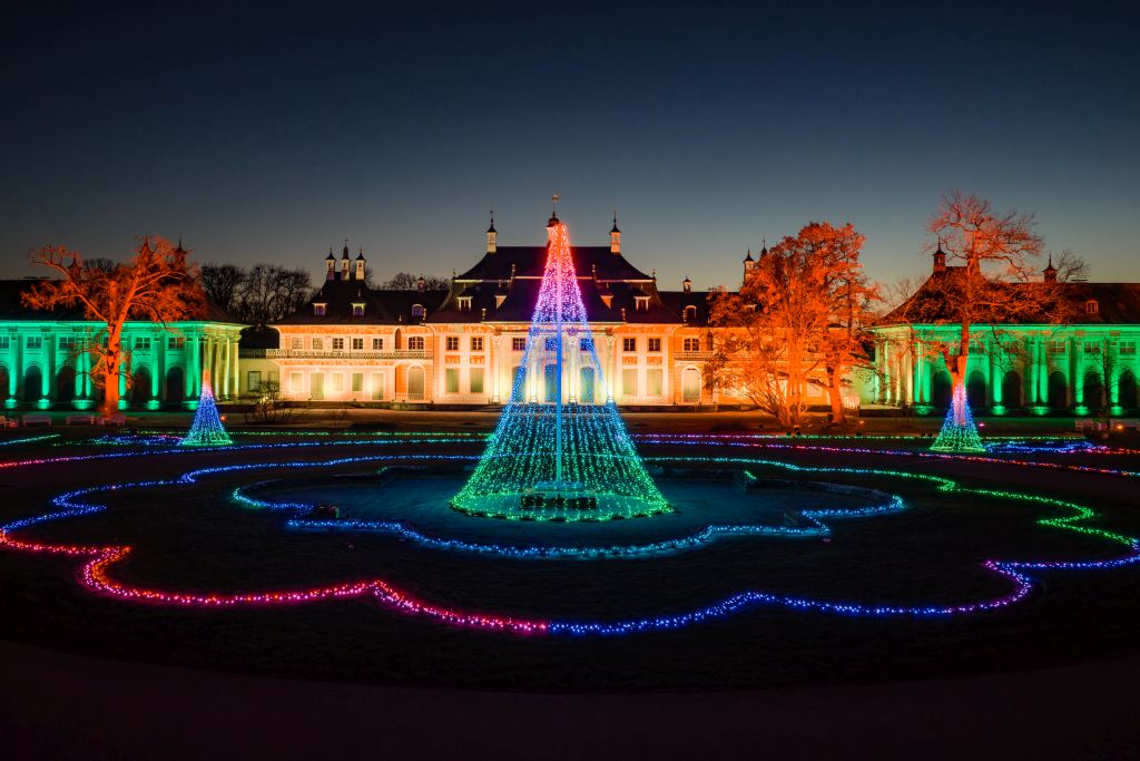 Ein großes historisches Gebäude ist nachts beleuchtet. Auf dem Rasen sind bunte Lichter in Mustern und Kegelformen angeordnet.