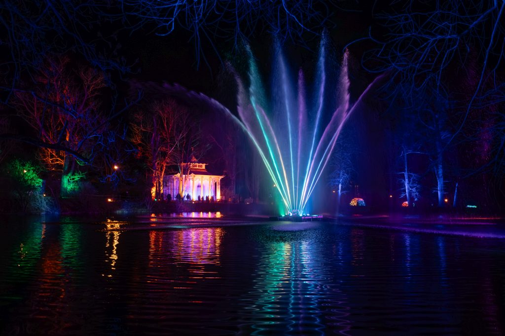 Beleuchtete Wasserspiele bei Nacht spiegeln sich in einem Teich. Im Hintergrund sind ein beleuchtetes Gebäude und Bäume zu sehen.