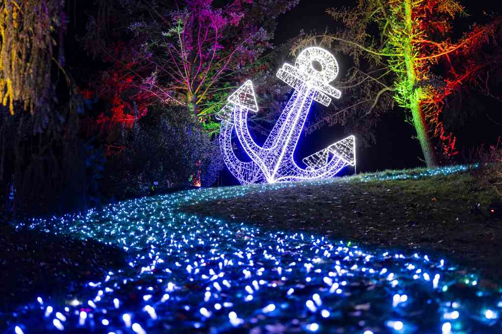 A large anchor made of white lights stands in a meadow. Trees and ground are decorated with blue and colorful lights.