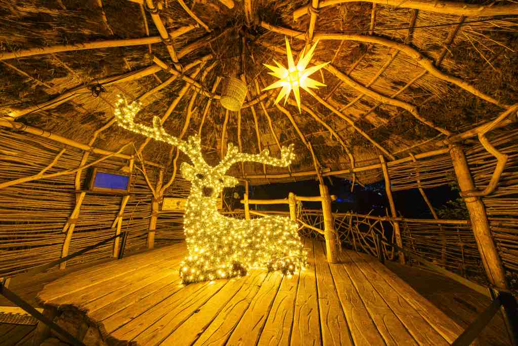 Eine Hirschskulptur mit gelben Lichterketten steht unter einem Reetdach. Darüber hängt ein sternförmiges Licht.