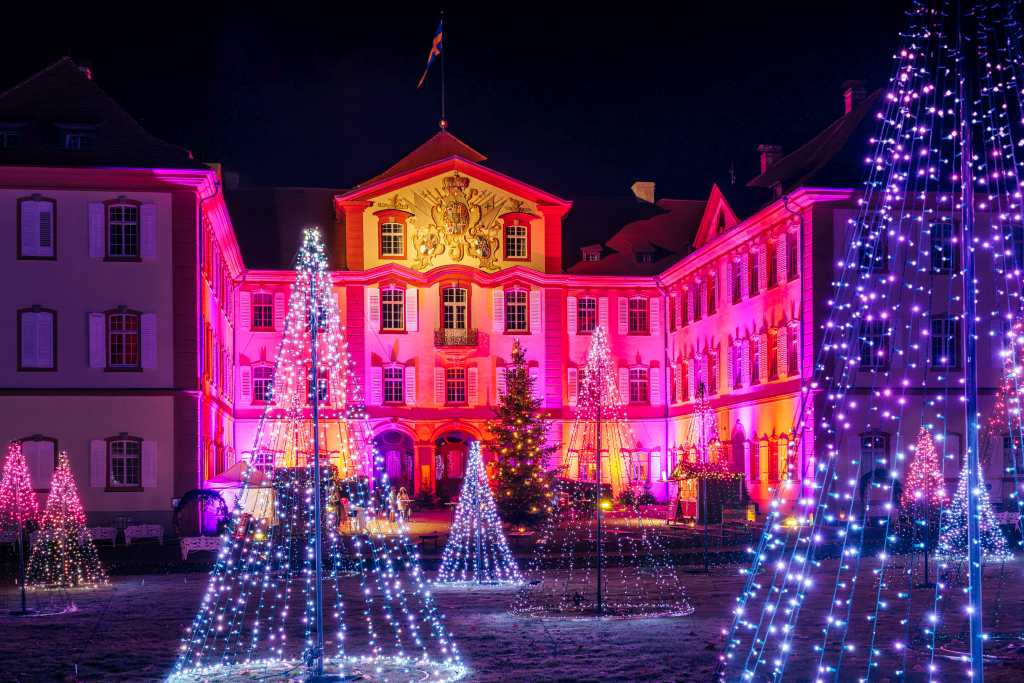 Ein großes Gebäude ist nachts rosa beleuchtet. Im Vordergrund stehen mehrere kegelförmige Lichtinstallationen.