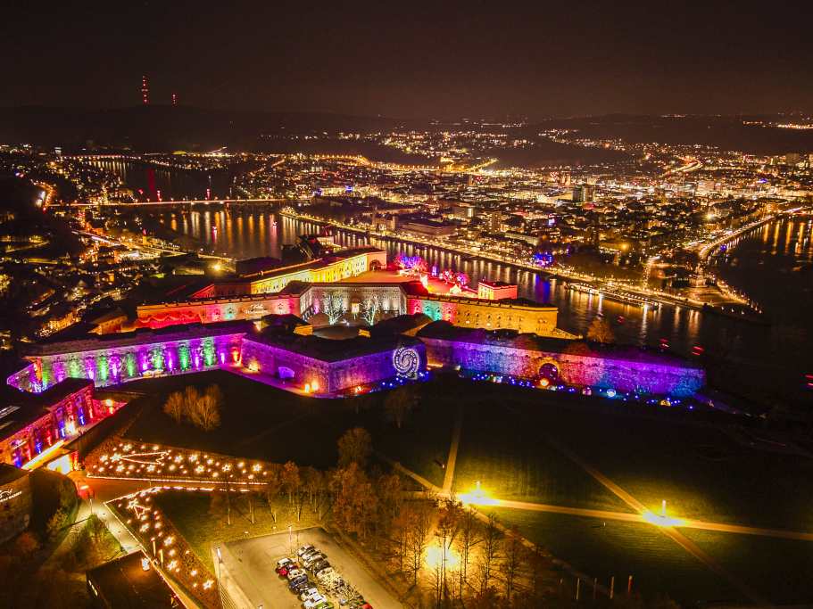 Luftaufnahme einer beleuchteten Festung und Stadt an einem Fluss bei Nacht. Bunte Lichter heben die Mauern der Festung hervor.