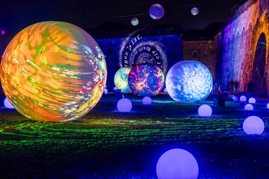 Große, leuchtende Kugeln mit verschiedenen Farben und Mustern stehen nachts im Freien. Um sie herum liegen kleinere, leuchtende Kugeln auf dem Rasen.