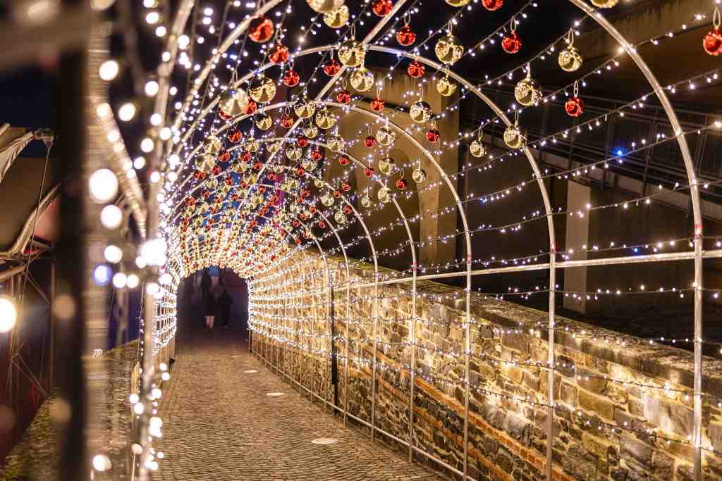 Ein Tunnel aus Metallbögen überspannt einen Pflasterweg. Er ist mit weißen Lichterketten und roten sowie goldenen Kugeln geschmückt.