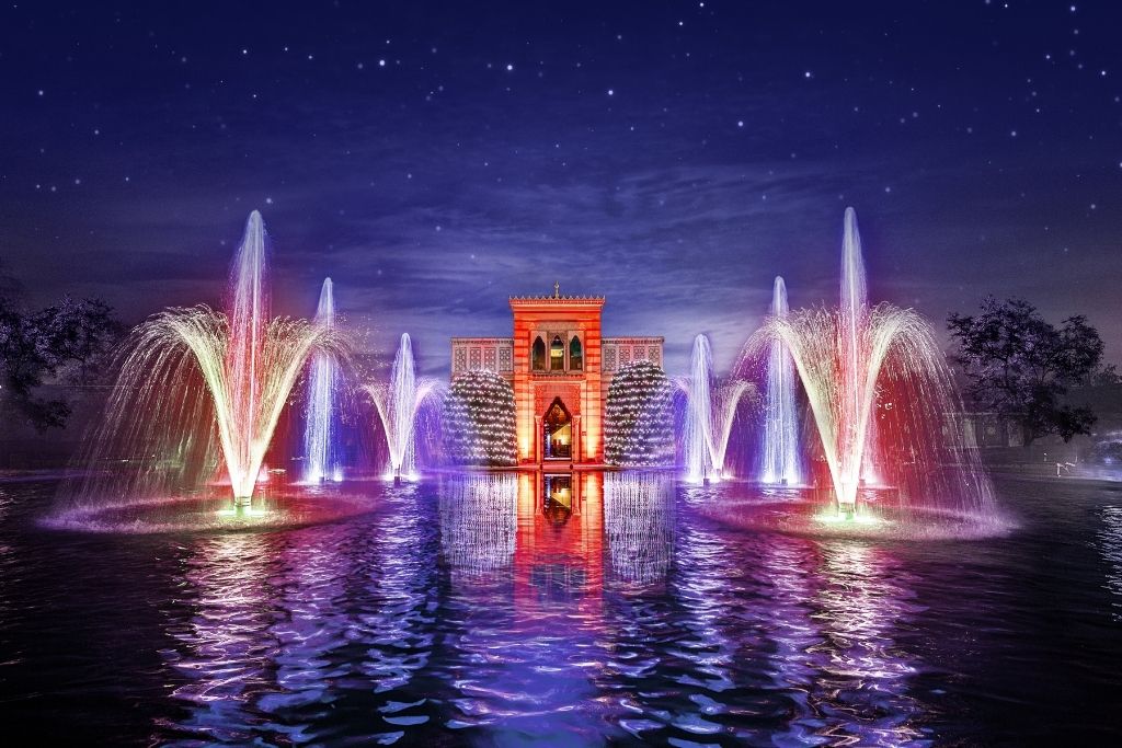 An illuminated fountain and a historic building are reflected in the water at night. You can see colorful lights and stars.
