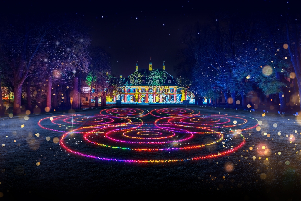 Ein großes Gebäude ist nachts bunt beleuchtet. Auf dem Rasen davor sind kreisförmige Lichtmuster zu sehen.