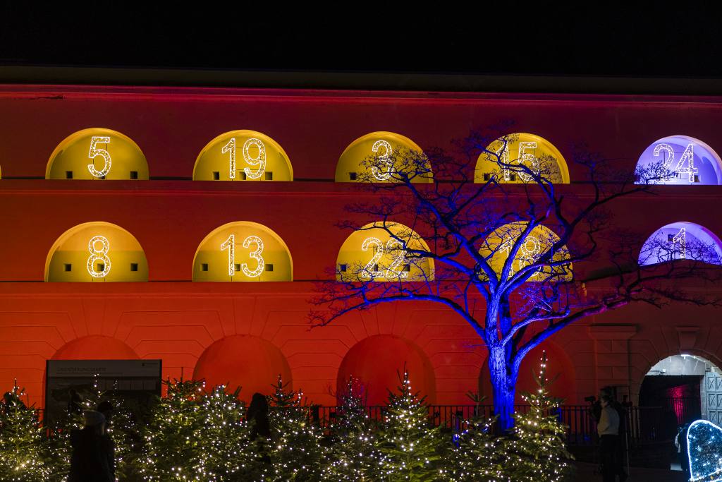 Ein Gebäude mit einer gelb und lila beleuchteten Fassade und nummerierten Fenstern. Im Vordergrund ein Baum mit Lichtern.