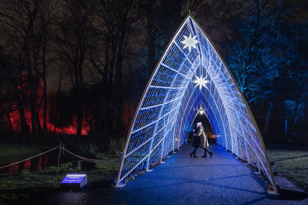Zwei Personen gehen nachts durch einen beleuchteten, dreieckigen Tunnel in einem Park mit farbig angestrahlten Bäumen.